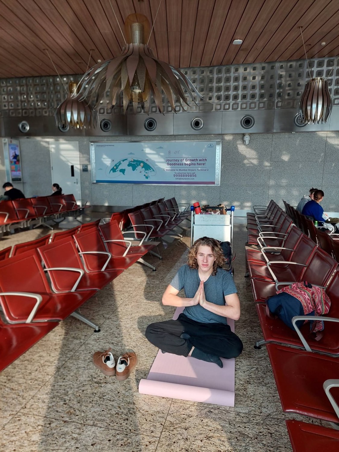 The artist sits cross-legged with palms together on a mat between rows of red seats in an airport terminal.