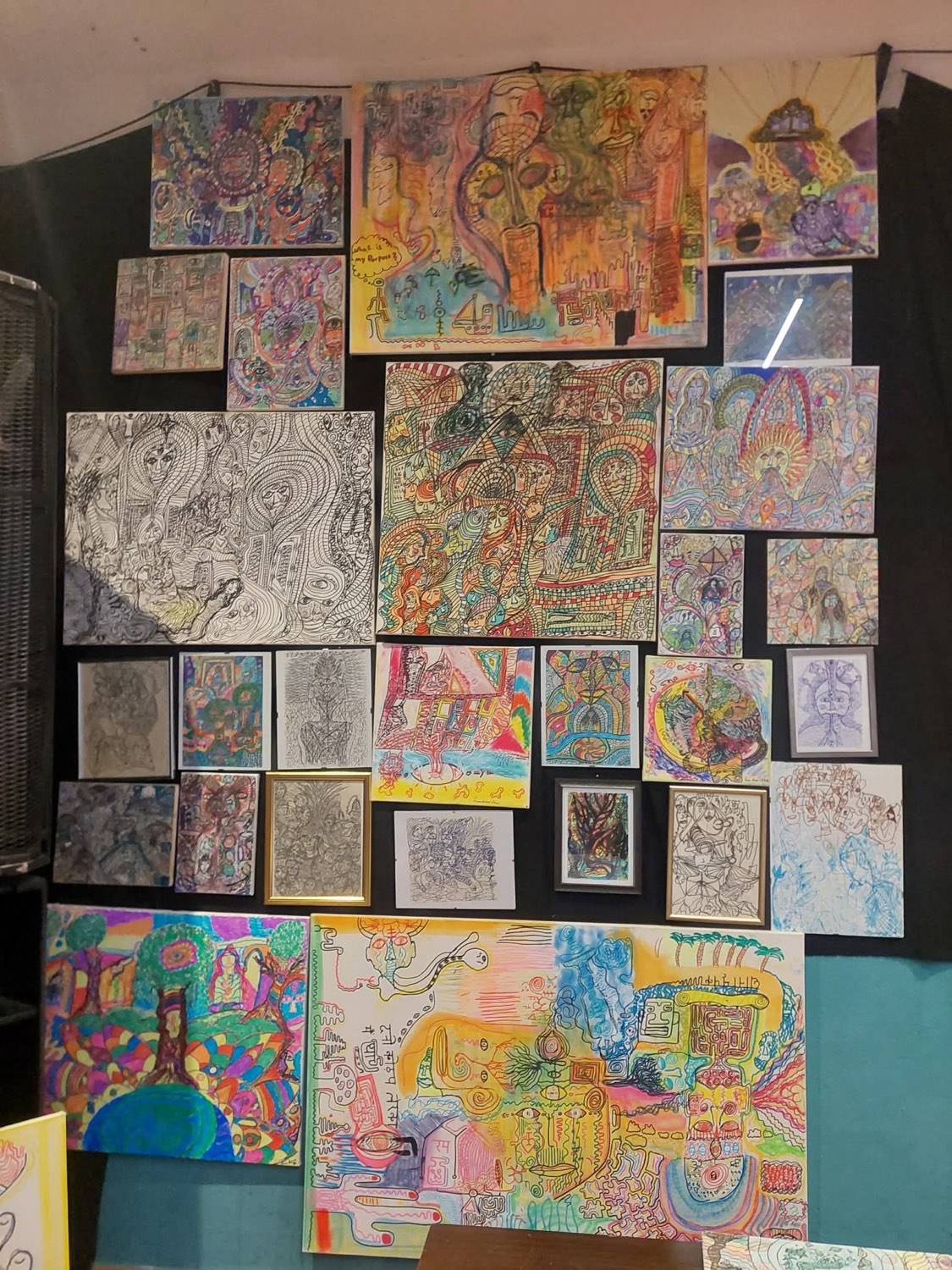 A dense wall of the artist’s works arranged edge to edge — colourful, densely patterned drawings alongside fine black-ink pieces.