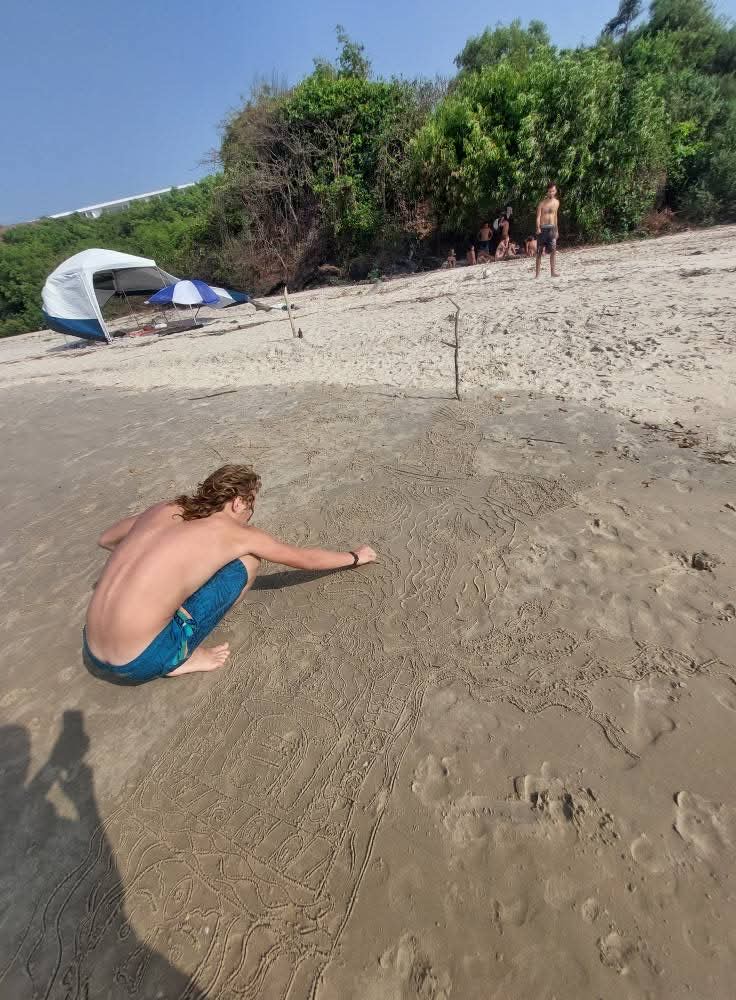 The artist crouches barefoot on a sunlit beach, drawing a large, intricate design into the sand with his hand; a tent and an umbrella stand near the treeline behind him.