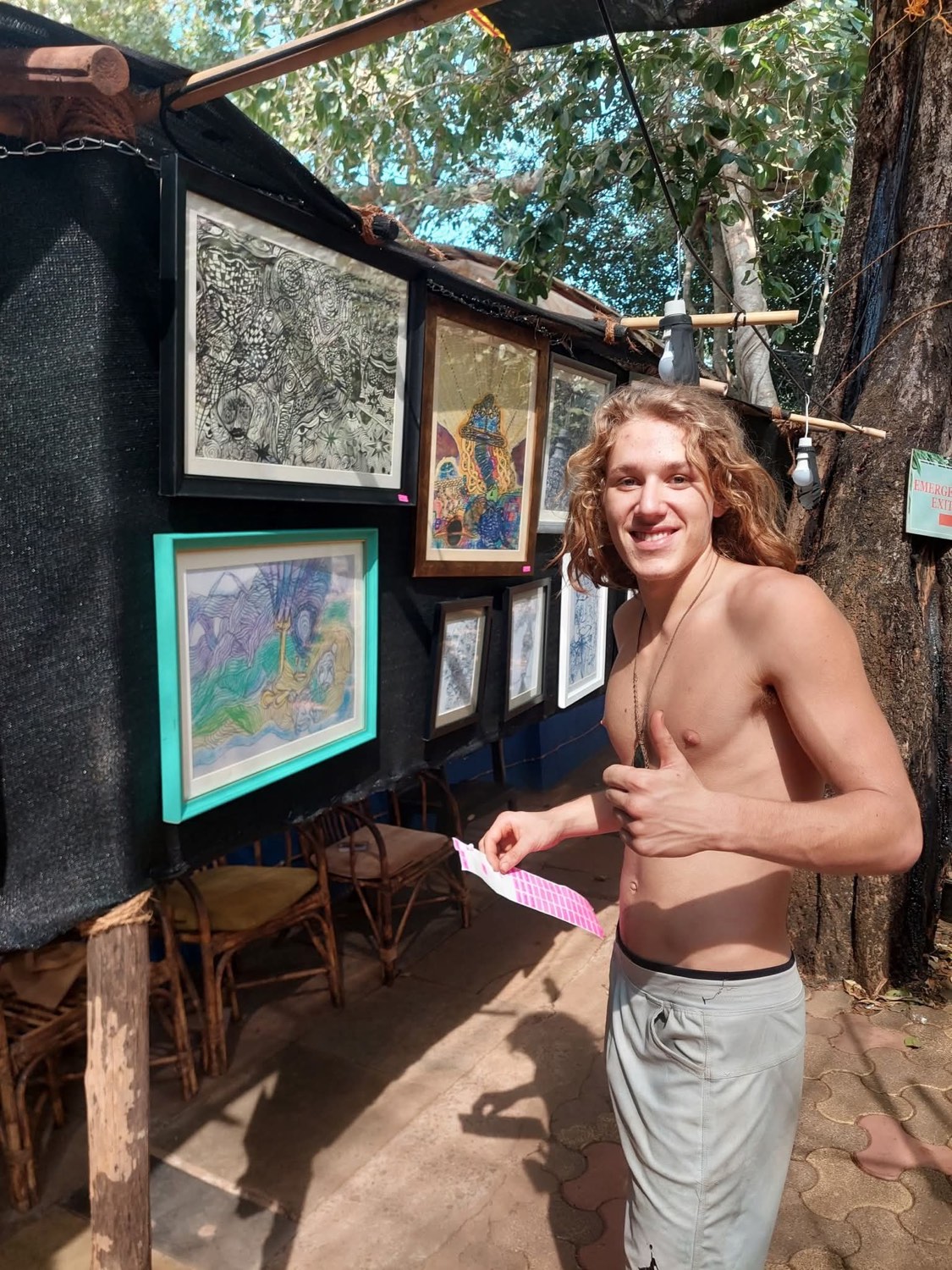 The artist smiles and gives a thumbs-up beside framed works displayed on a black outdoor wall among trees.
