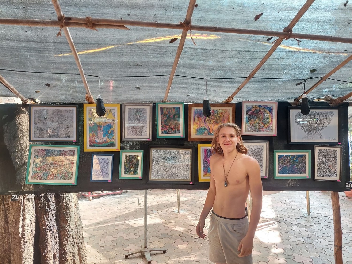 The artist stands beside a large outdoor wall hung with many framed drawings and paintings under a bamboo canopy.