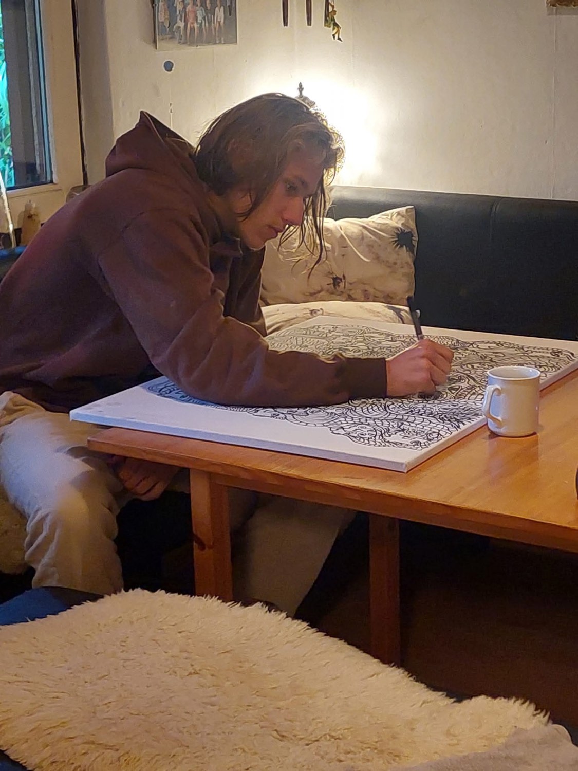 In a hooded jacket, the artist leans over a large sheet indoors, drawing dense black-ink linework by lamplight with a mug beside him.